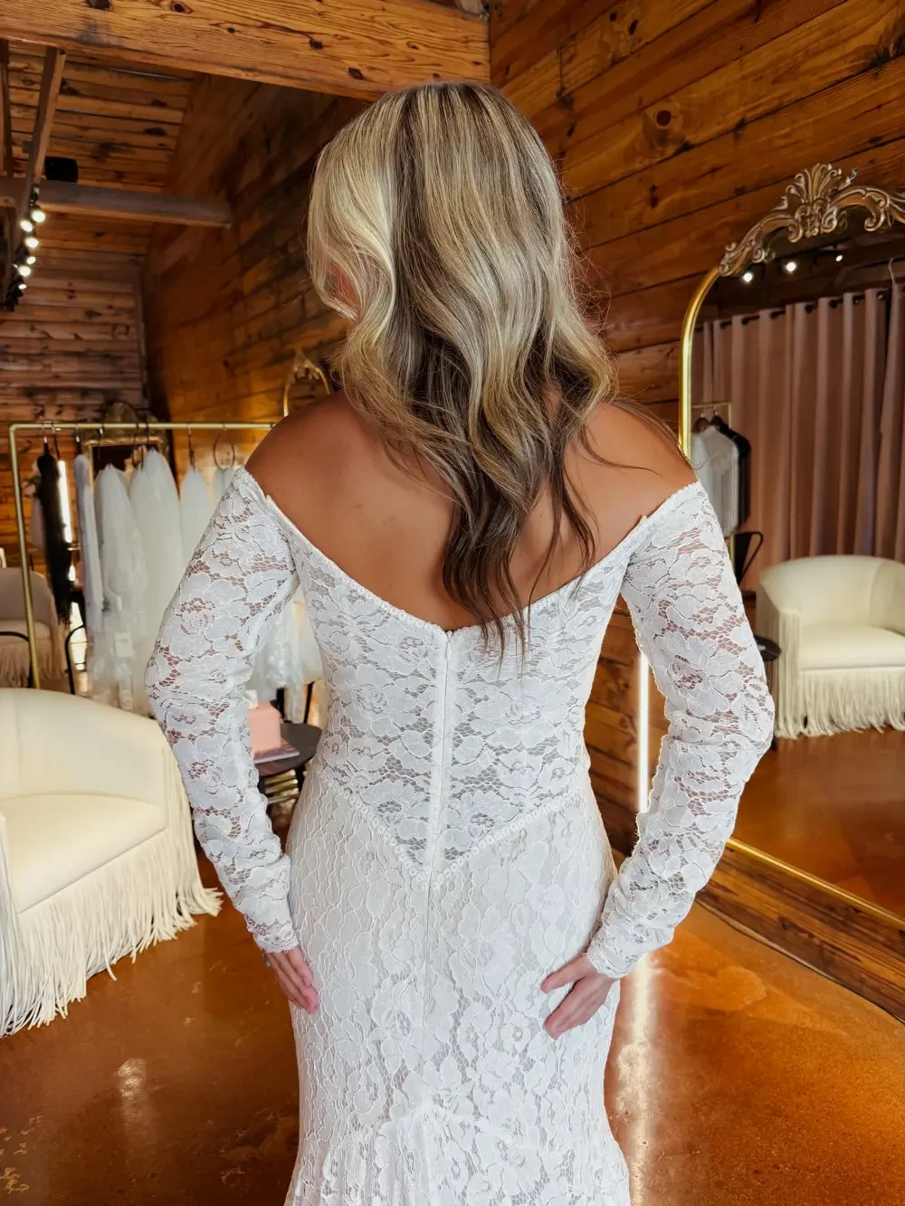 A woman wearing a white lace dress is seen from the back, with her long hair styled in loose waves. The background features a rustic wooden interior, mirrors, and bridal gowns hanging on a rack.