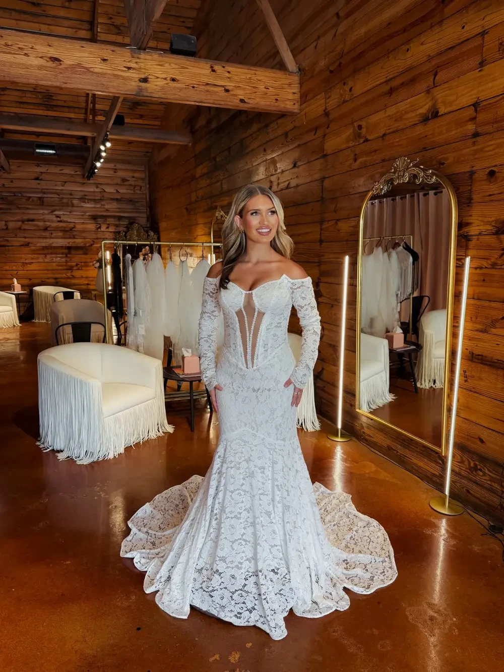 A woman in a white lace wedding dress stands in a rustic room with wooden walls, surrounded by bridal gowns and a large mirror.