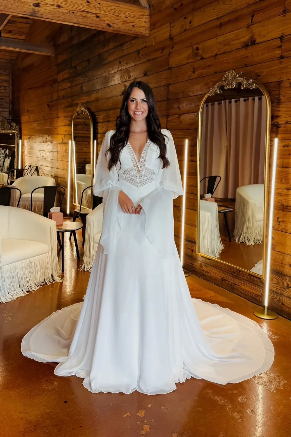 A woman in a white wedding dress standing in a room with wooden walls and mirrors, smiling at the camera.